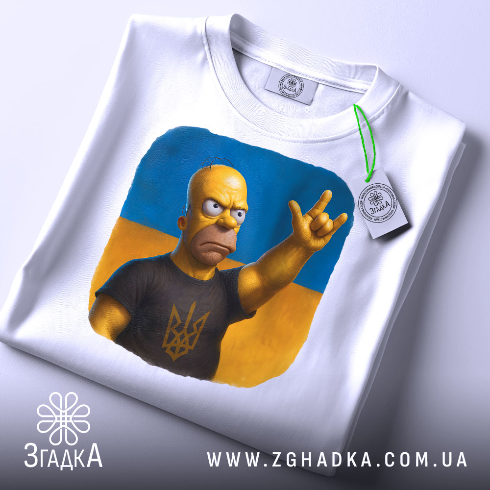 Футболка гомер з тризубом, біла, зображена на столі, фон світло-сірий, принт з гомером у чорній футболці з тризубом, на футболці є етикетка з зеленим шнурком.