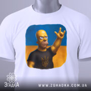 Футболка гомер з тризубом, біла, зображена на столі, фон світло-сірий, принт з гомером у чорній футболці з тризубом, на футболці є етикетка з зеленим шнурком.