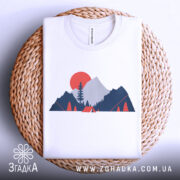 Футболка гори кемпінг, біла, складена на кошику, фон світлий, принт з горами, деревом та сонцем у червоному кольорі.