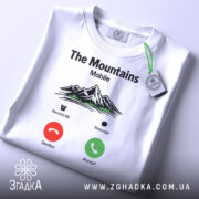 Футболка гори на зв’язку, біла, складена на столі, фон світло-сірий, принт з горами, текстом 'The Mountains Mobile' та іконками дзвінка, з зеленим ярликом.