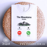 Футболка гори на зв’язку, біла, складена на плетеній тарілці, фон світло-сірий, принт з горами, текстом 'The Mountains Mobile' та іконками дзвінка.