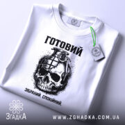 Футболка готовий до дій, біла, зображена зверху, на ній підвішений етикет, фон світло-сірий, принт з черепом та гранатою, текст "Готовий. Зібраний. Спокійний." під малюнком.
