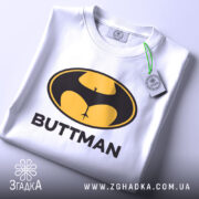 Футболка гумор буттмен, біла, складена на поверхні, фон світло-сірий, принт з жовтим і чорним логотипом у формі кажана з написом 'BUTTMAN', на комірці видна етикетка.