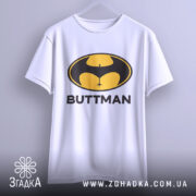 Футболка гумор буттмен, біла, підвішена на вішалці, фон світло-сірий, принт з жовтим і чорним логотипом у формі кажана з написом 'BUTTMAN'.