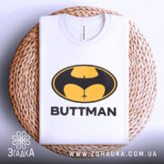 Футболка гумор буттмен, біла, складена на плетеній кошику, фон світло-сірий, принт з жовтим і чорним логотипом у формі кажана з написом 'BUTTMAN', видна етикетка на комірці.