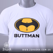 Футболка гумор буттмен, біла, складена на поверхні, фон світло-сірий, принт з жовтим і чорним логотипом у формі кажана з написом 'BUTTMAN', на комірці видна етикетка.