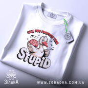 Футболка гусак з характером, біла, складена на столі, фон світло-сірий, принт з гусаком, який тримає мозок, текст "HEY, YOU DROPPED THIS STUPID" в яскравих кольорах, з етикеткою на комірці.