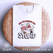 Футболка гусак з характером, біла, складена на плетеному кошику, фон білий, принт з гусаком, який тримає мозок, текст "HEY, YOU DROPPED THIS STUPID" в яскравих кольорах.