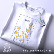 Футболка гуси гумор, біла, складена на поверхні, фон світло-сірий, принт з жовтими гусячими головами та текстом 'what?' у верхній частині, з зеленим ярликом.