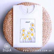 Футболка гуси гумор, біла, складена на плетеній кошику, фон світло-сірий, принт з жовтими гусячими головами та текстом 'what?' у верхній частині.