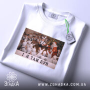 Футболка і я там був. Біла футболка, складена вдвічі, з принтом, на якому діти у зайчих костюмах спостерігають за Дідом Морозом. Принт яскравий, фон святковий. Текст на принті чорний: 'I Я ТАМ БУВ'. На футболці є етикетка з зеленим шнурком.