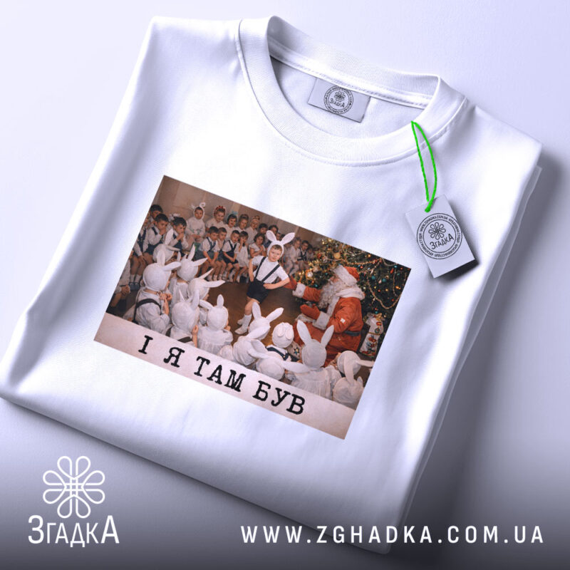 Футболка і я там був. Біла футболка, складена вдвічі, з принтом, на якому діти у зайчих костюмах спостерігають за Дідом Морозом. Принт яскравий, фон святковий. Текст на принті чорний: 'I Я ТАМ БУВ'. На футболці є етикетка з зеленим шнурком.