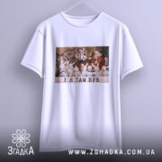 Футболка і я там був. Біла футболка, підвішена на вішалці, з принтом, що зображує дітей у зайчих костюмах, які дивляться на Діда Мороза. Принт кольоровий, фон святковий. Текст на принті чорний: 'I Я ТАМ БУВ'.