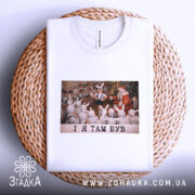 Футболка і я там був. Біла футболка, складена на плетеній тарілці, з принтом, на якому діти у зайчих костюмах спостерігають за Дідом Морозом. Принт яскравий, фон святковий. Текст на принті чорний: 'I Я ТАМ БУВ'.