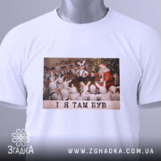 Футболка і я там був. Біла футболка, складена вдвічі, з принтом, на якому діти у зайчих костюмах спостерігають за Дідом Морозом. Принт яскравий, фон святковий. Текст на принті чорний: 'I Я ТАМ БУВ'. На футболці є етикетка з зеленим шнурком.