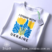 Футболка I Stand Ukraine білого кольору, складена на поверхні, фон світло-сірий, принт з жовтими квітами та синіми елементами, а також напис 'UKRAINE', на футболці є етикетка з зеленим шнурком.