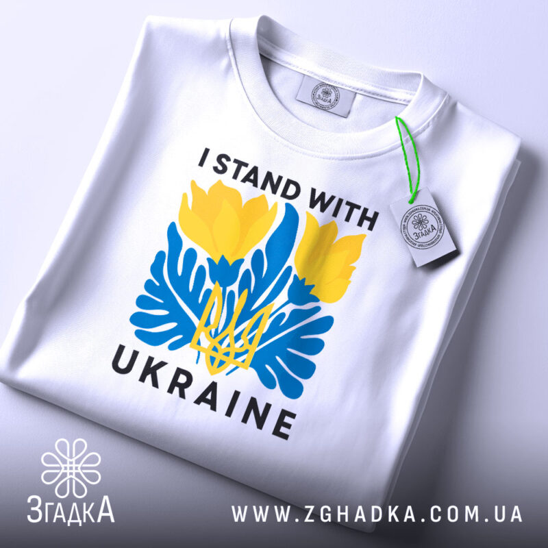 Футболка I Stand Ukraine білого кольору, складена на поверхні, фон світло-сірий, принт з жовтими квітами та синіми елементами, а також напис 'UKRAINE', на футболці є етикетка з зеленим шнурком.