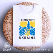 Футболка I Stand Ukraine білого кольору, складена на круглій кошику, фон білий, принт з жовтими квітами та синіми елементами, а також напис 'UKRAINE'.
