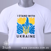 Футболка I Stand Ukraine білого кольору, складена на поверхні, фон світло-сірий, принт з жовтими квітами та синіми елементами, а також напис 'UKRAINE', на футболці є етикетка з зеленим шнурком.