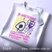 Футболка я анестезіолог, білого кольору, зображена з верхнього ракурсу, на ній прикріплений етикетка, фон світло-сірий, принт містить малюнок стетоскопа та текст у яскравих кольорах: жовтому, рожевому та фіолетовому.