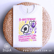 Футболка я анестезіолог, білого кольору, зображена на плетеній кошику, фон білий, принт містить малюнок стетоскопа та текст у яскравих кольорах: жовтому, рожевому та фіолетовому.