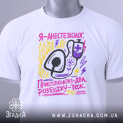 Футболка я анестезіолог, білого кольору, зображена з верхнього ракурсу, на ній прикріплений етикетка, фон світло-сірий, принт містить малюнок стетоскопа та текст у яскравих кольорах: жовтому, рожевому та фіолетовому.