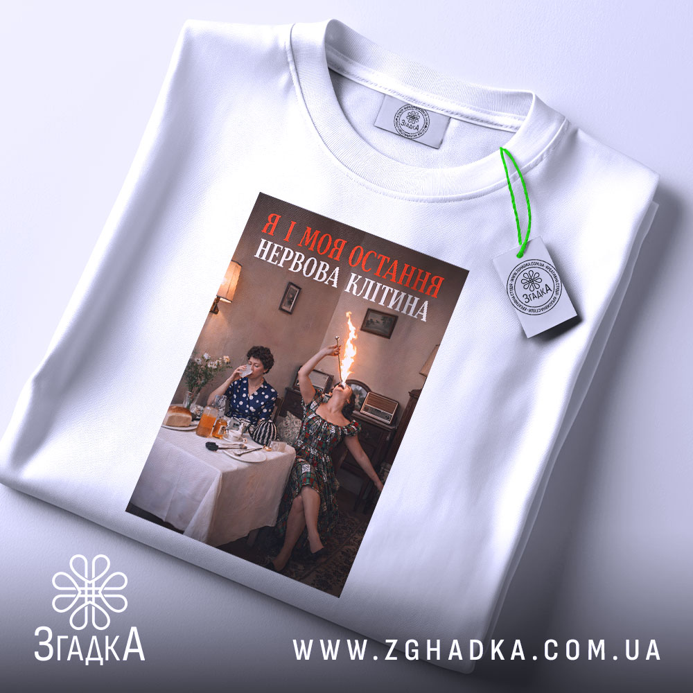 Футболка я і нерви. Біла футболка, складена на столі, з принтом, що зображує двох жінок, одна з яких підносить пляшку з вогнем, фон - затишна кімната з меблями та столом, заставленим їжею.