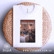 Футболка я і нерви. Біла футболка, складена на плетеному кошику, з принтом, що зображує двох жінок за столом, одна з яких тримає пляшку з вогнем, фон - затишна обстановка з елементами декору.