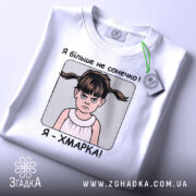 Футболка я хмарка, біла, складена на столі, фон світло-сірий, принт з дівчинкою з двома хвостиками та написом 'Я більше не сонечко! Я - ХМАРКА!', на футболці є етикетка з зеленим шнурком.