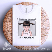 Футболка я хмарка, біла, складена на плетеній кошику, фон світло-сірий, принт з дівчинкою з двома хвостиками та написом 'Я більше не сонечко! Я - ХМАРКА!'.