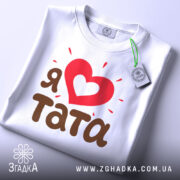 Футболка я люблю тата, білого кольору, складена на поверхні з легким нахилом, фон світло-сірий, принт з червоним серцем та коричневим текстом 'тата', на футболці є зелений ярлик.