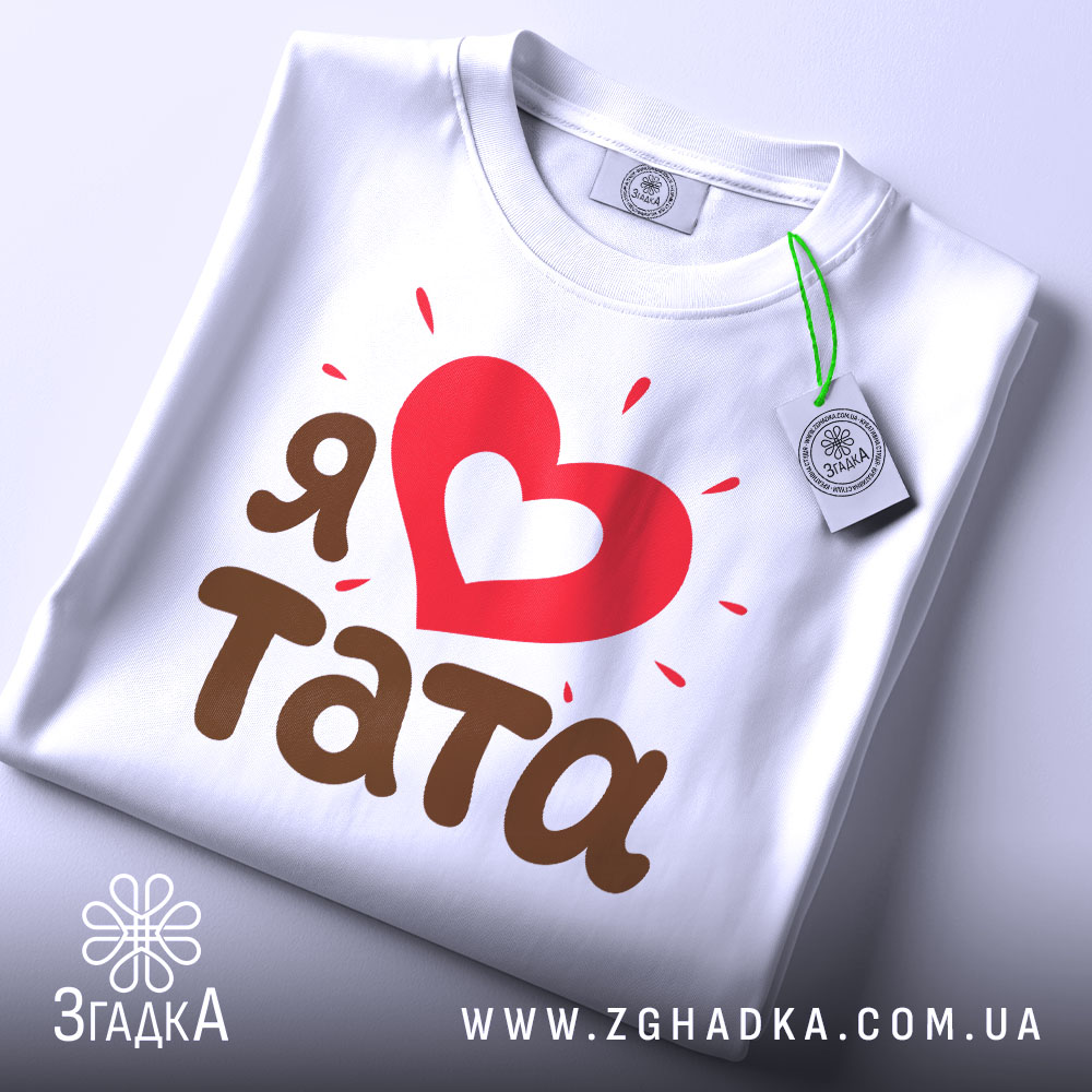Футболка я люблю тата, білого кольору, складена на поверхні з легким нахилом, фон світло-сірий, принт з червоним серцем та коричневим текстом 'тата', на футболці є зелений ярлик.