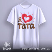 Футболка я люблю тата, білого кольору, підвішена на вішалці, фон світло-сірий, принт з червоним серцем та коричневим текстом 'тата', виглядає стильно та сучасно.