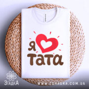 Футболка я люблю тата, білого кольору, складена на плетеній кошику, фон світло-сірий, принт з червоним серцем та коричневим текстом 'тата', виглядає затишно та привабливо.