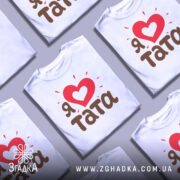 Футболка я люблю тата, білого кольору, зображена на манекені з блискучого металу, фон світло-сірий, принт містить червоне серце з білим серцем всередині та коричневий текст 'тата' з червоними спалахами навколо.