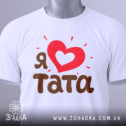 Футболка я люблю тата, білого кольору, складена на поверхні з легким нахилом, фон світло-сірий, принт з червоним серцем та коричневим текстом 'тата', на футболці є зелений ярлик.