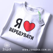 Футболка я люблю вередувати, біла, складена на плоскій поверхні, фон світлий, принт з червоним сердечком і чорним текстом, на комірці є етикетка.