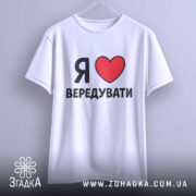 Футболка я люблю вередувати, біла, підвішена на вішалці, фон світло-сірий, принт з червоним сердечком і чорним текстом, видно округлі краї рукавів.