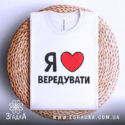 Футболка я люблю вередувати, біла, складена на плоскій поверхні з плетеним кошиком на фоні, принт з червоним сердечком і чорним текстом.