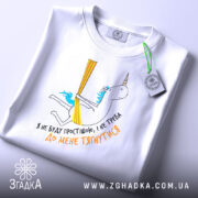 Футболка я не буду простішою. Біла футболка, складена на поверхні, з принтом єдинорога. На футболці видно етикетку з зеленим шнурком, принт кольоровий, з текстом українською мовою.
