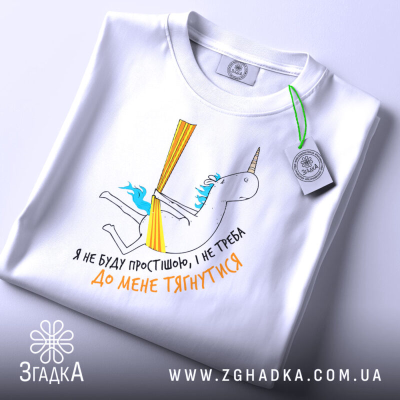 Футболка я не буду простішою. Біла футболка, складена на поверхні, з принтом єдинорога. На футболці видно етикетку з зеленим шнурком, принт кольоровий, з текстом українською мовою.