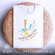 Футболка я не буду простішою. Біла футболка, складена на плетеній основі, з принтом єдинорога. Принт кольоровий, з текстом українською мовою, видно етикетку на комірці.