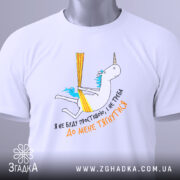 Футболка я не буду простішою. Біла футболка, складена на поверхні, з принтом єдинорога. На футболці видно етикетку з зеленим шнурком, принт кольоровий, з текстом українською мовою.