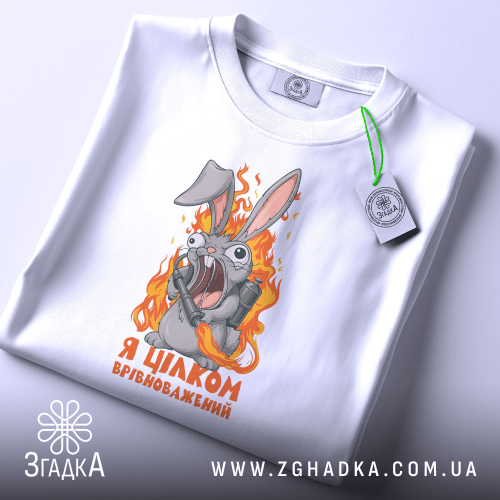Футболка я цілком врівноважений, білого кольору, складена на столі, фон світло-сірий, принт з кроликом, який виглядає емоційно, тримаючи гранату, текст на футболці чорного кольору, з зеленим ярликом.