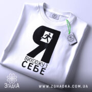 Футболка я виходжу з себе. Біла футболка складена, з чорним принтом, зображення чоловіка, що біжить, видно чітко. На футболці є етикетка з зеленим шнурком. Фон світло-сірий.