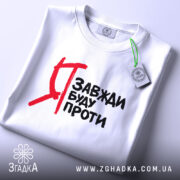 Футболка я завжди буду проти, білого кольору, зображена зверху, на столі, фон світлий, принт чорного кольору з червоними акцентами, на футболці видно етикетку з зеленим шнурком.