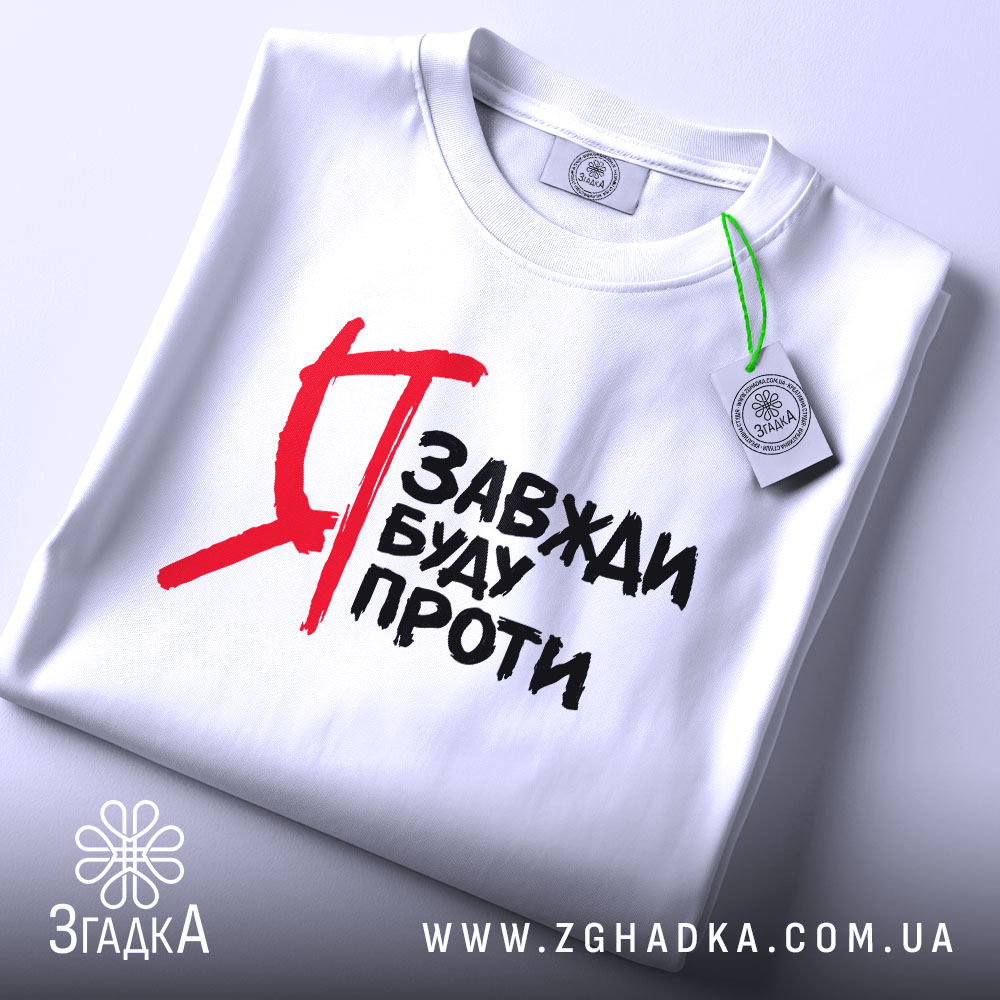 Футболка я завжди буду проти, білого кольору, зображена зверху, на столі, фон світлий, принт чорного кольору з червоними акцентами, на футболці видно етикетку з зеленим шнурком.