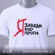 Футболка я завжди буду проти, білого кольору, зображена зверху, на столі, фон світлий, принт чорного кольору з червоними акцентами, на футболці видно етикетку з зеленим шнурком.