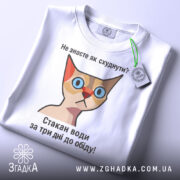 Футболка як швидко схуднути. Біла футболка, складена на поверхні, з етикеткою зеленого кольору. Принт з кішкою та текстом: 'Не знаєте як схуднути? Стакан води за три дні до обіду!' Фон світлий, що підкреслює футболку.