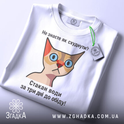 Футболка як швидко схуднути. Біла футболка, складена на поверхні, з етикеткою зеленого кольору. Принт з кішкою та текстом: 'Не знаєте як схуднути? Стакан води за три дні до обіду!' Фон світлий, що підкреслює футболку.